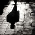 Blurry shadow and silhouette of a man standing in the night on wet city street sidewalk with water reflection in black and white